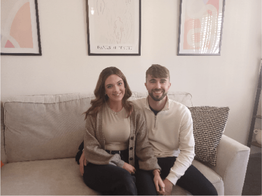 Young couple in a home, both sat down on a white/cream coloured couch